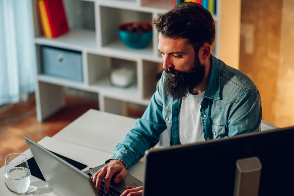 A young focused bearded writer is sitting at home and typing an article on a laptop. A writer in smart casual is sitting at home and typing a report on a laptop while working remotely.