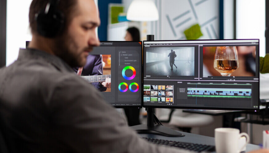 Videographer with headset working with footage and sound on pc with two displays, taking notes while editing customer video. Man processing film montage sitting in creative startup agency office.